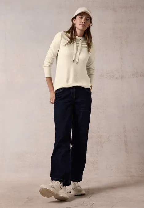 Frau mit cremefarbenem Pullover, dunkler Hose, Turnschuhen und Kappe steht vor hellem Hintergrund.